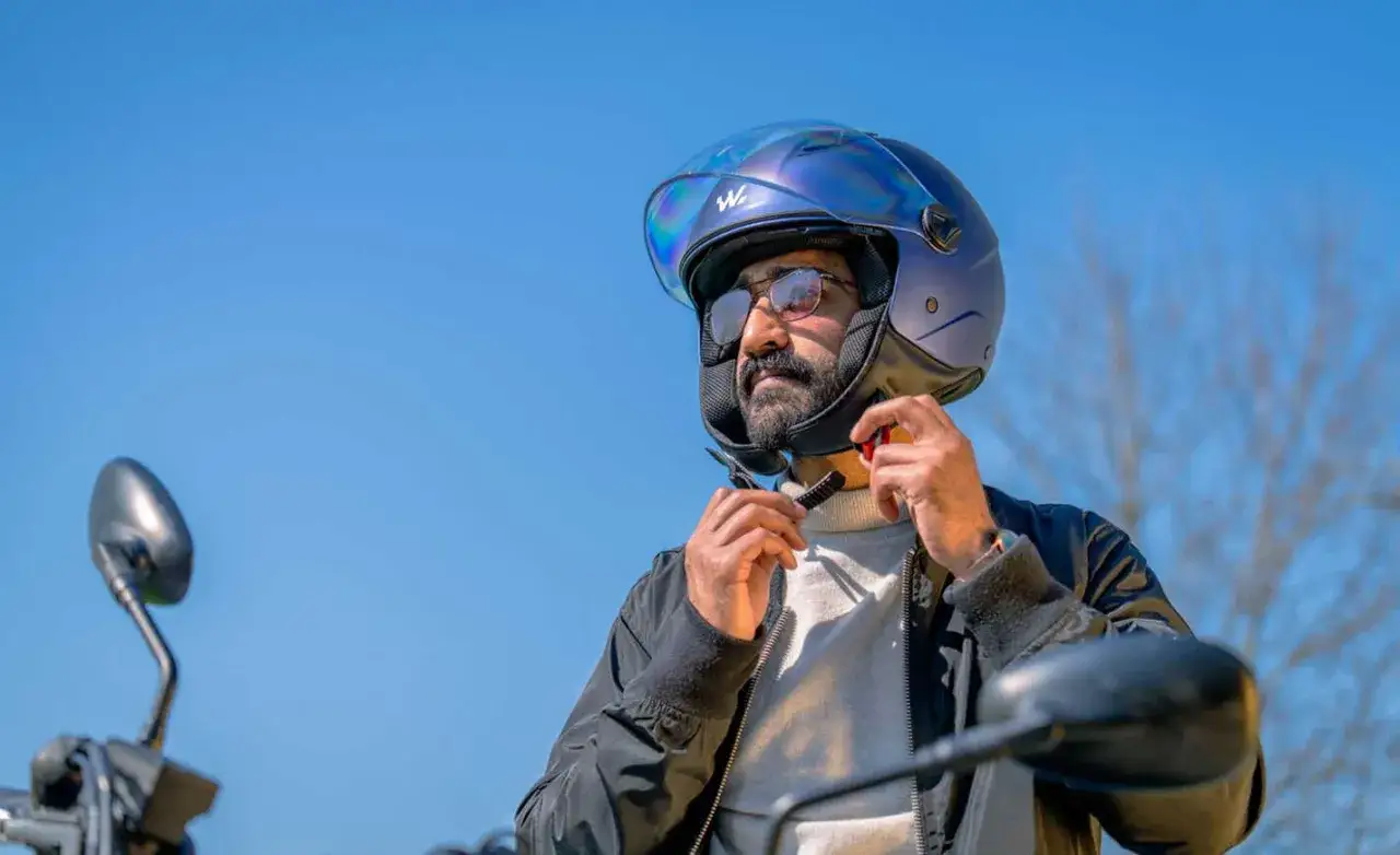 Uomo con barba e occhiali indossa un casco stretto blu iridescente, pronto per un giro in moto sotto il cielo limpido.