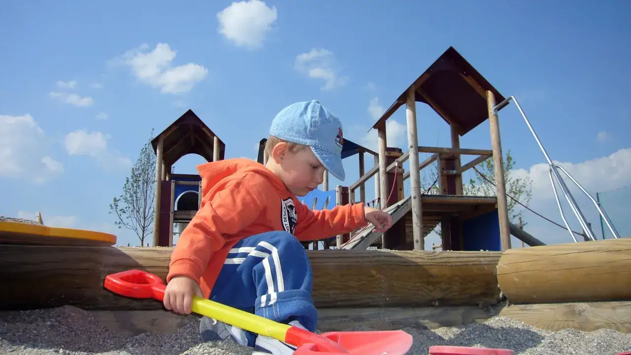 Ein Junge mit blauer Kappe und orangefarbener Jacke spielt allein im Sandkasten. Er wirkt vertieft, fast wie ein typisches Einzelkind, das sich in seiner eigenen Welt verliert.