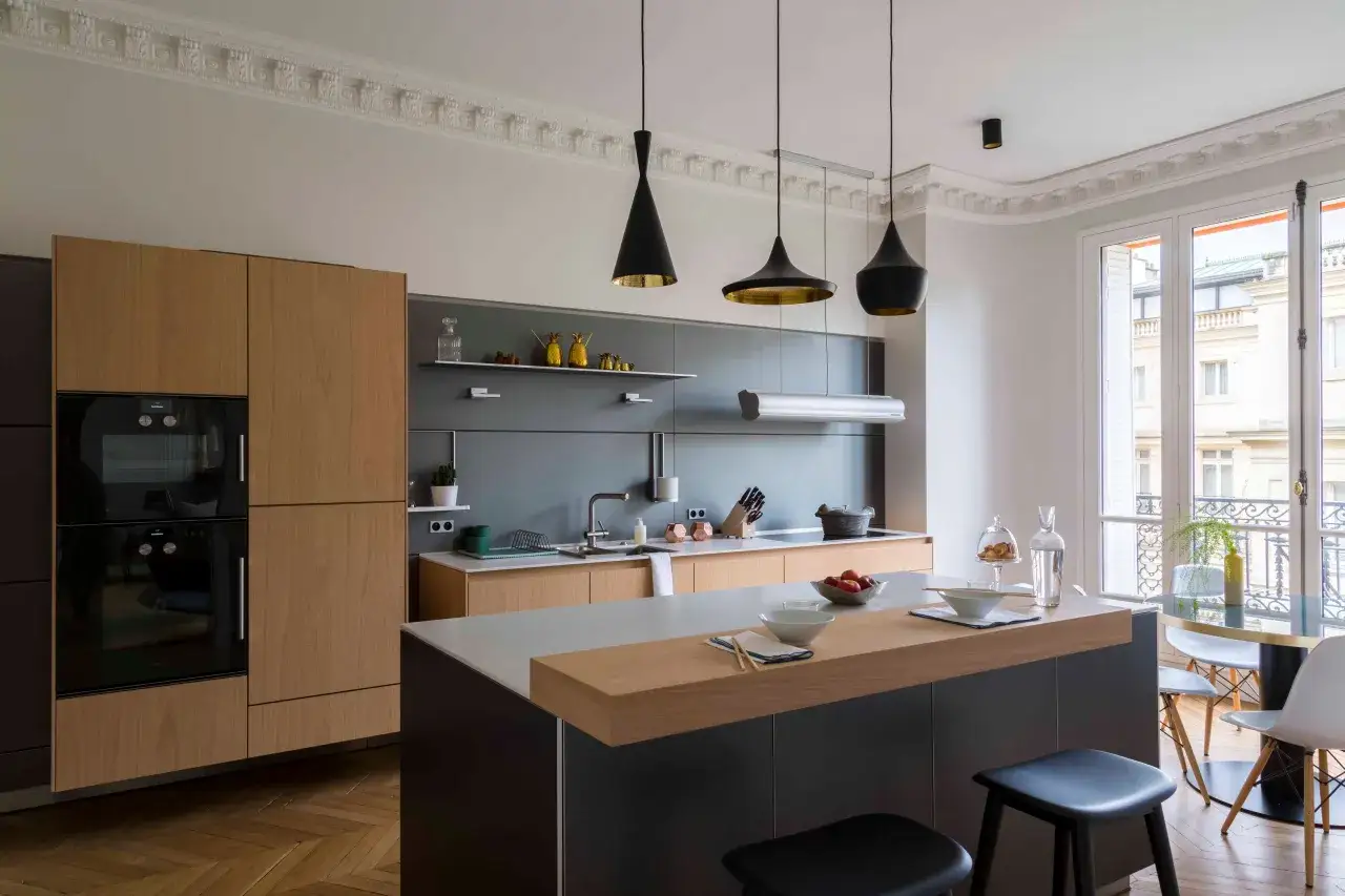 Cuisine moderne avec &icirc;lot central, placards en bois clair et carrelage marron fonc&eacute; au sol. Luminaires noirs suspendus.