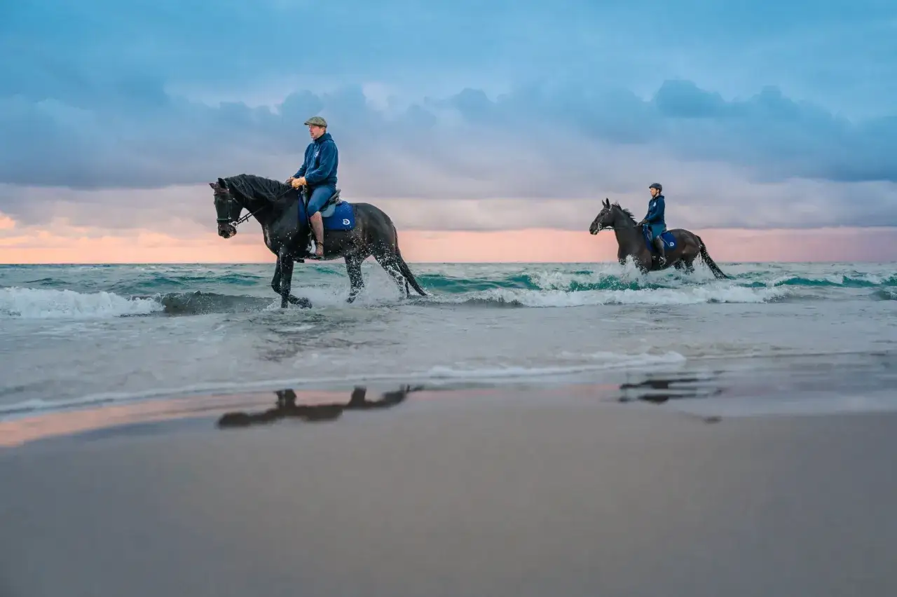 Zwei Reiter auf schwarzen Pferden durchqueren das Meer am Strand. Das Wasser spritzt um die Hufe, während der Himmel in sanften Pastelltönen leuchtet.