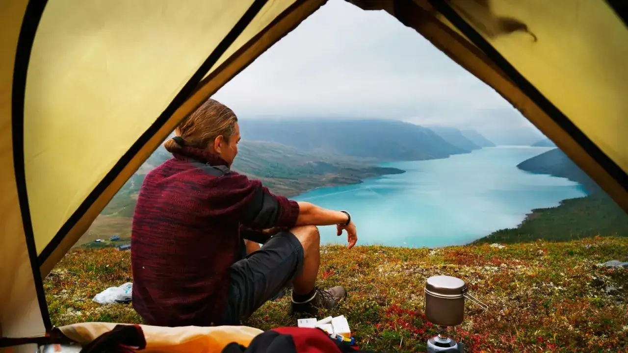 En person sitter vid t&auml;lt&ouml;ppningen och blickar ut &ouml;ver en turkos sj&ouml; omgiven av berg. En perfekt vy fr&aring;n en av Norges m&aring;nga campingplatser.