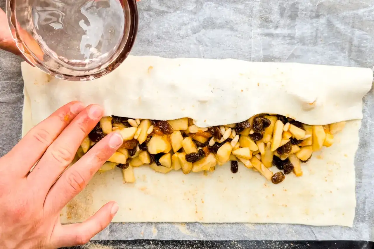 Mani che chiudono uno strudel con ripieno di mele, uvetta e pinoli, preparato con pasta sfoglia o frolla.