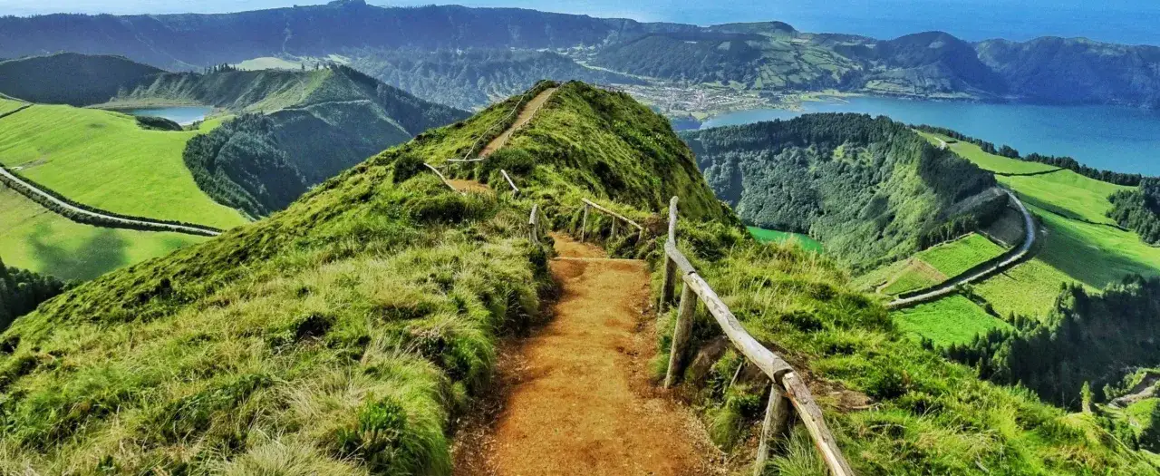 A scenic hiking trail winds along a lush green ridge, offering breathtaking views of volcanic lakes and rolling hills, perfect for hiking trips in Portugal.
