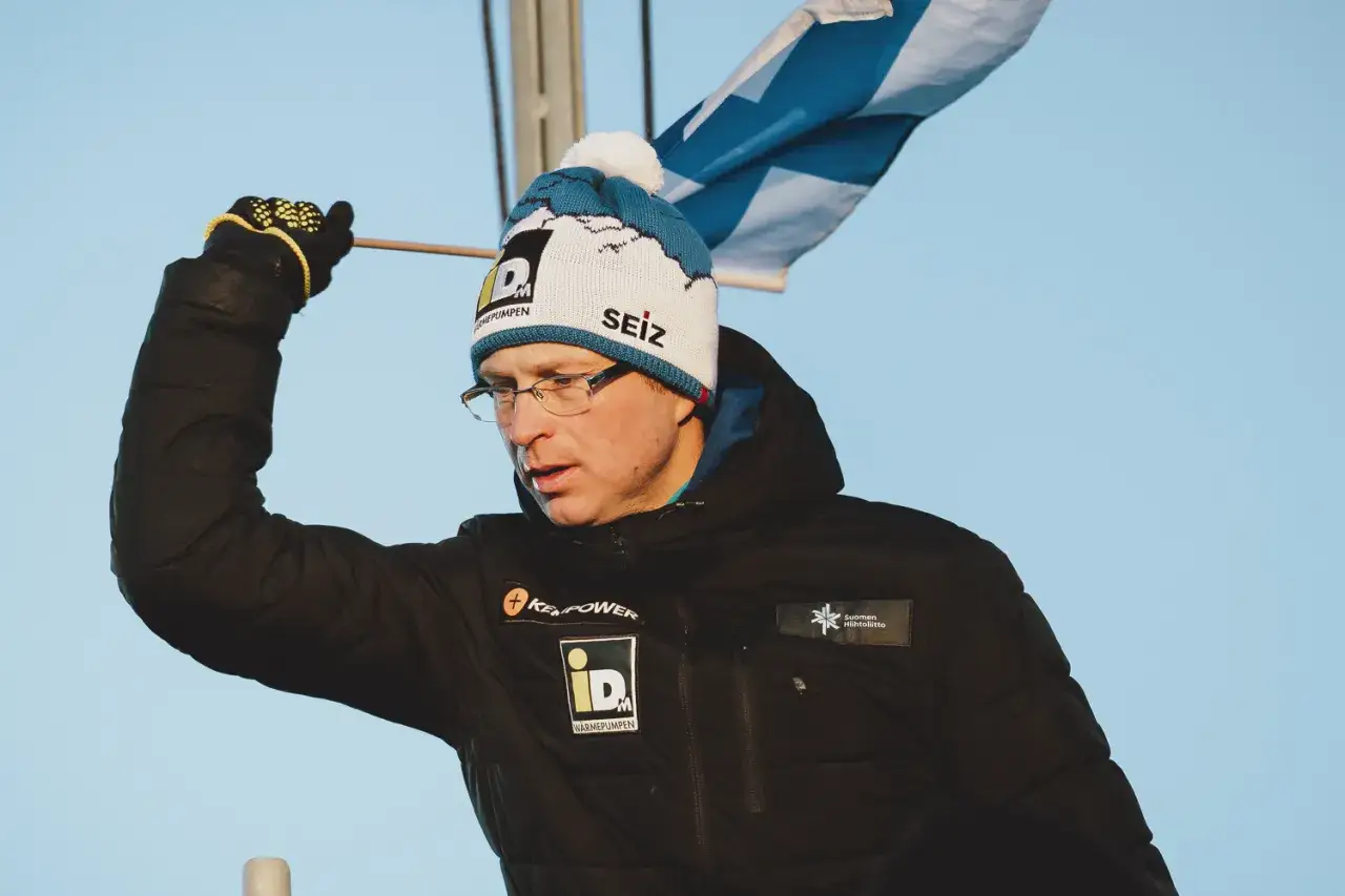 Igor Medved w czapce z pomponem i flagą Finlandii, ubrany w czarną kurtkę z logo.