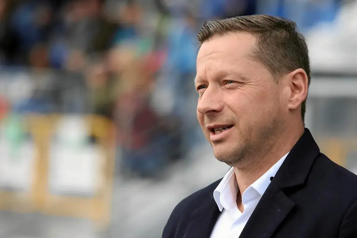 Dariusz Banasik, w czarnym garniturze i białej koszuli, z uwagą słucha, co dzieje się na tle rozmytego tłumu.