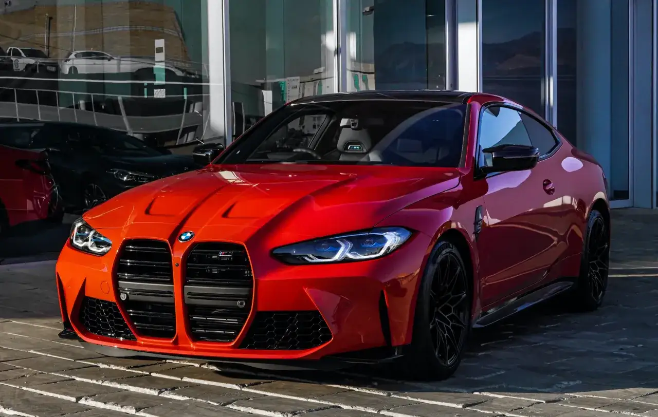 En r&ouml;d, sportig tysk bil, en BMW M4, st&aring;r parkerad framf&ouml;r en modern byggnad med stora glaspartier.