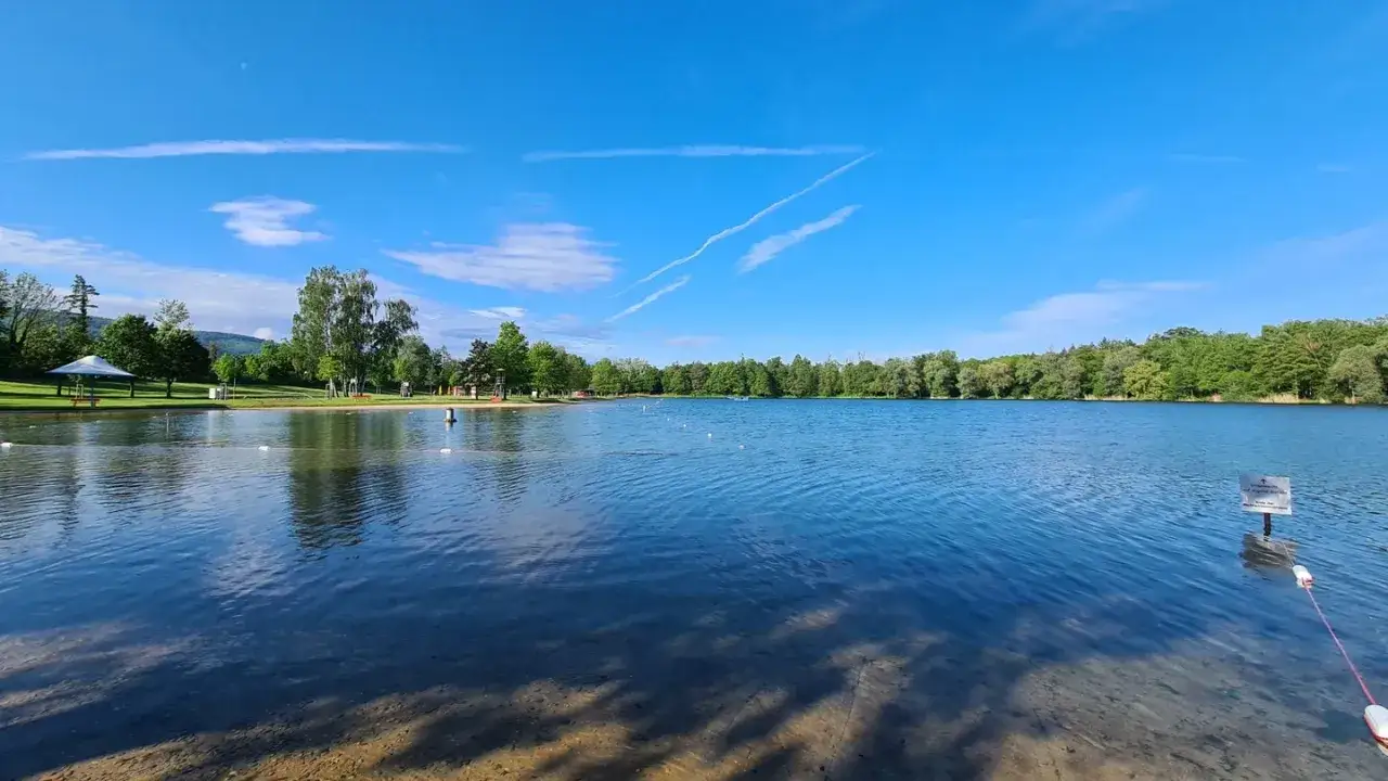 Ein idyllischer **Badesee im Schwarzwald** unter blauem Himmel. Gr&uuml;ne B&auml;ume s&auml;umen das Ufer, w&auml;hrend das klare Wasser zum Schwimmen einl&auml;dt.