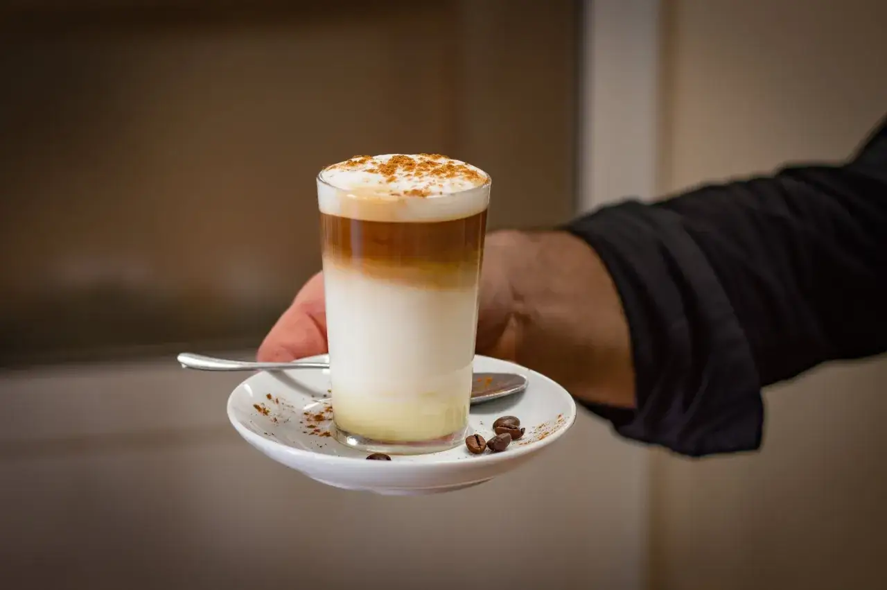 Dłoń podaje szklankę z warstwowym espresso macchiato, posypane cynamonem, na talerzyku z ziarnami kawy.