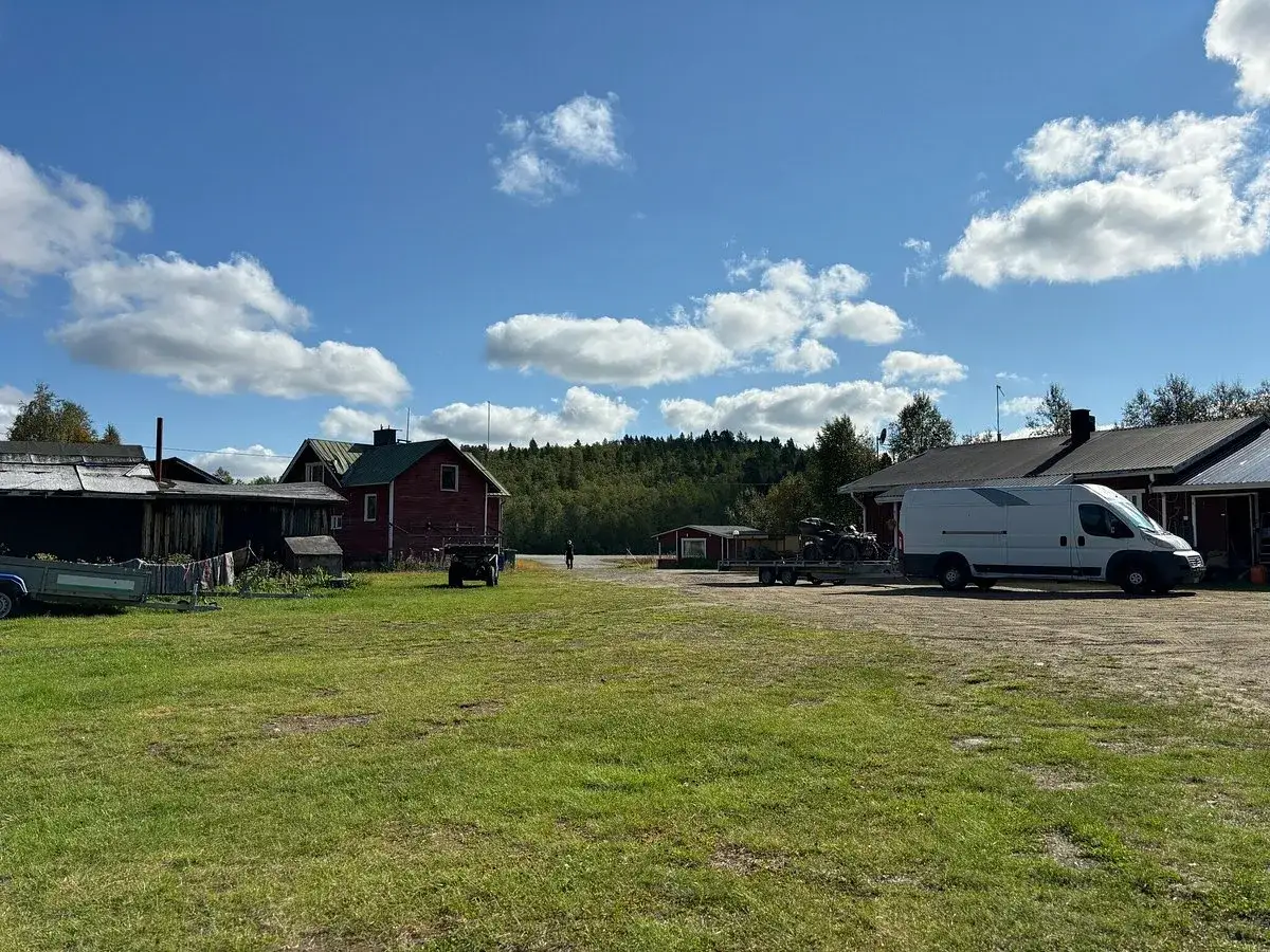 Karesuando camping: Röd stuga, vit skåpbil med släp och fyrhjuling på gräsplan under molnig himmel.