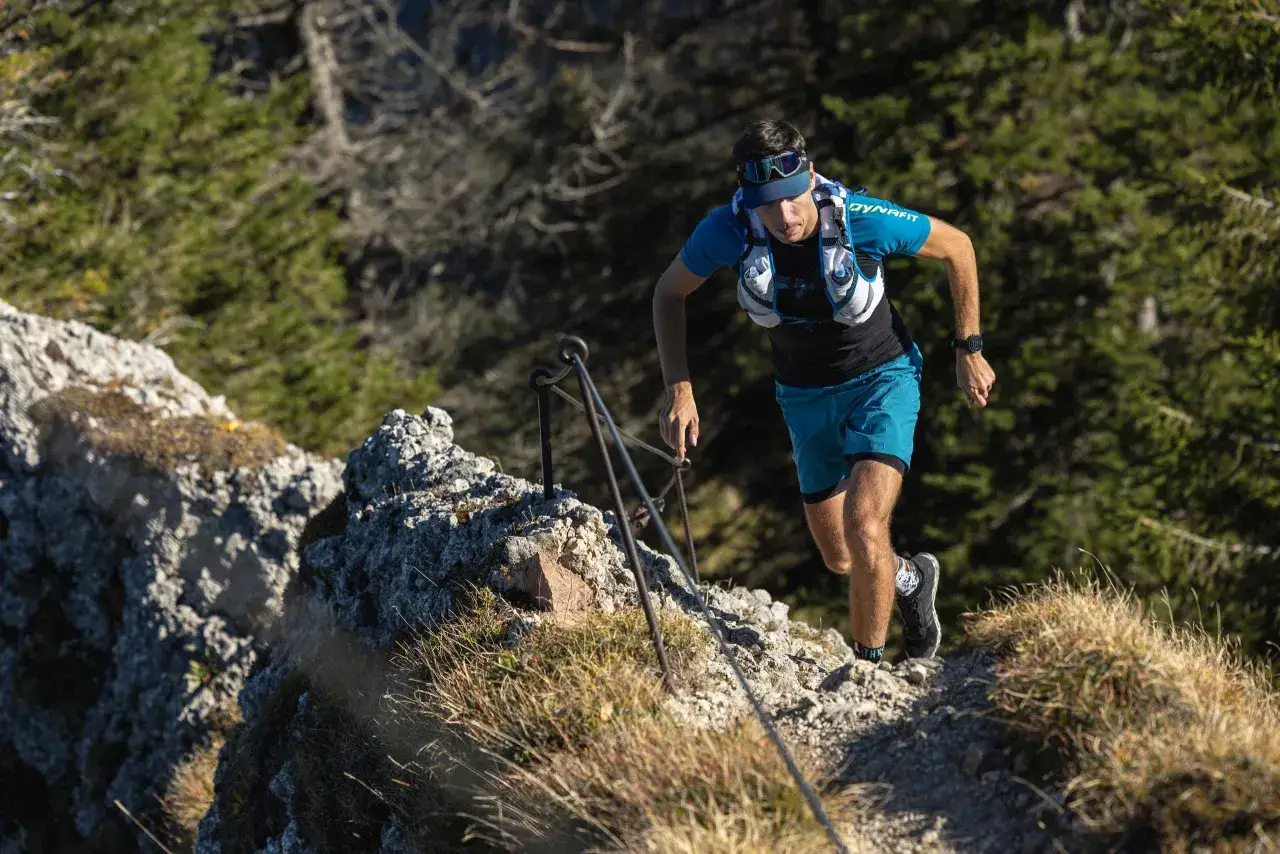 Ein Mann läuft auf einem steinigen Pfad bergauf, mit Blick auf die Zugspitze. Er trägt Sportkleidung und einen Laufrucksack.
