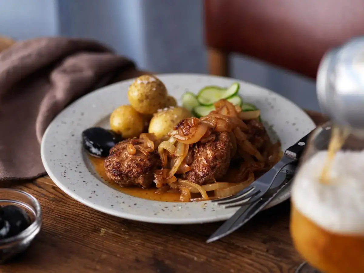 En tallrik med pannbiff, stekt l&ouml;k, kokt potatis, gurka och en m&ouml;rk plommon. Ett perfekt pannbiff recept f&ouml;r en mysig middag.