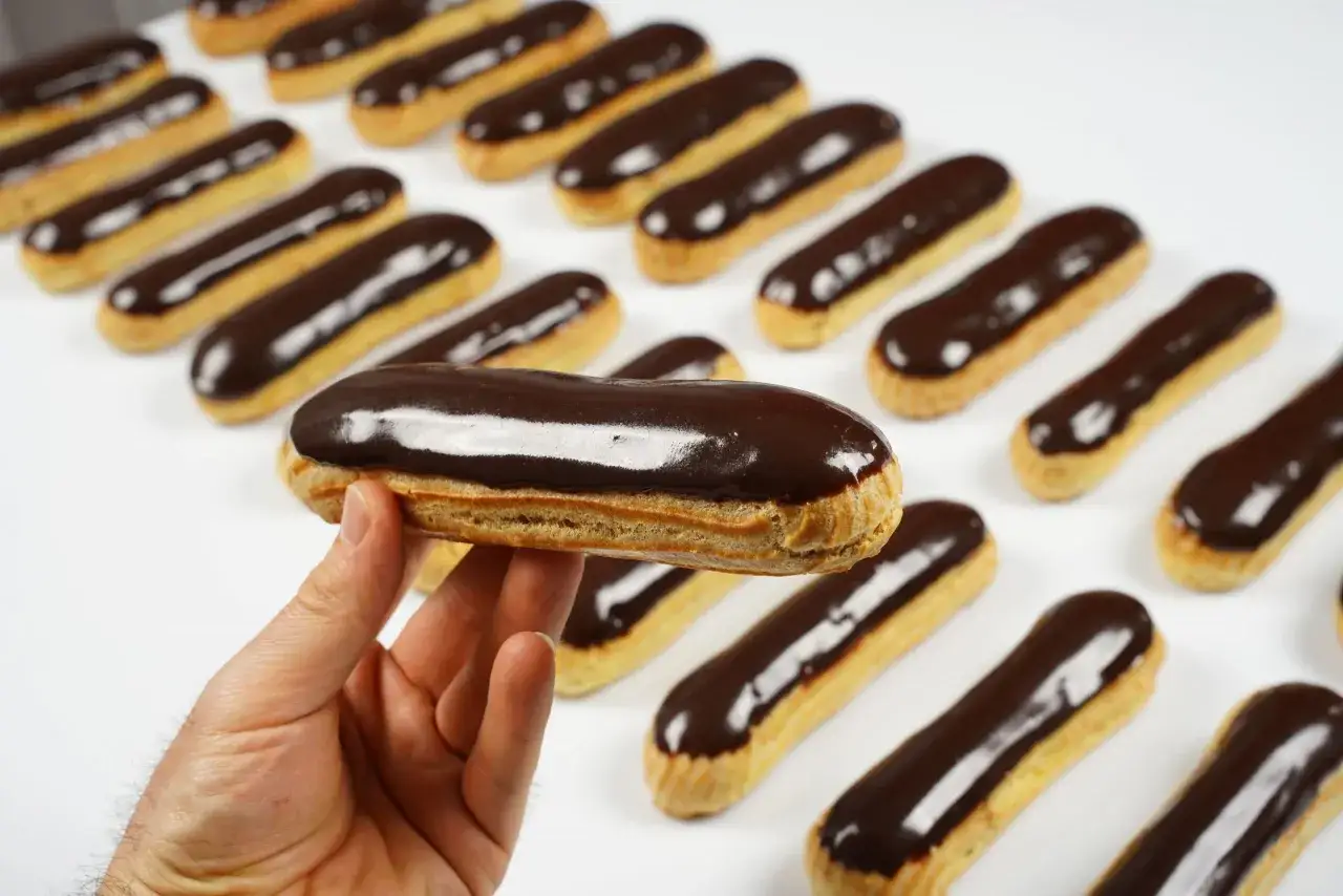 Une main tient un éclair au glaçage au chocolat brillant, avec une rangée d'autres éclairs en arrière-plan.