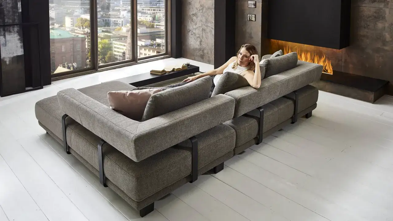 Ein großes, graues Sofa mitten im Raum, mit einer Frau, die sich darauf ausruht. Im Hintergrund ein Kamin und ein Fenster mit Stadtblick.