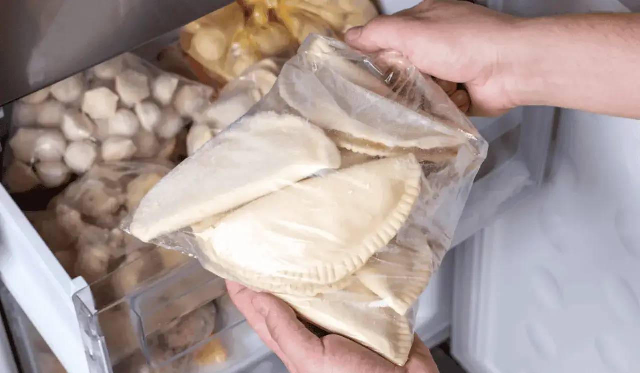 Ręce wyjmują z zamrażarki worek z surowymi pierogami. W zamrażarce widać też inne mrożone pierogi.
