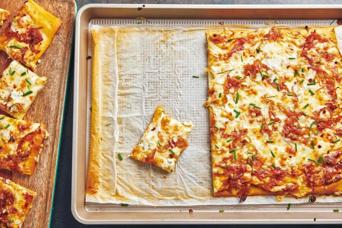 Rechteckige Pizza Formaggi mit geschmolzenem K&auml;se und Schnittlauch, daneben auf einem Holzbrett geschnittene St&uuml;cke.