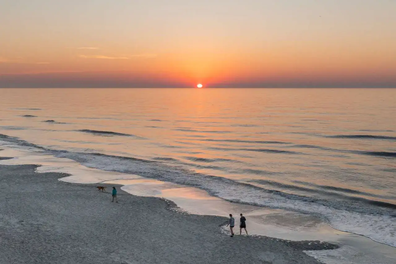 Zachód słońca na plaży, idealne miejsce, gdzie na wakacje w lutym. Dwie osoby spacerują z psem.