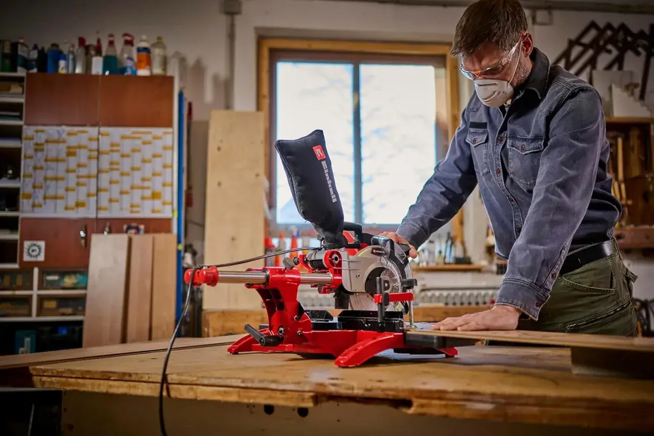 Un homme en masque et lunettes utilise une scie à onglet rouge pour couper du bois. Il est dans un atelier, prêt à choisir scie à onglet pour son projet.