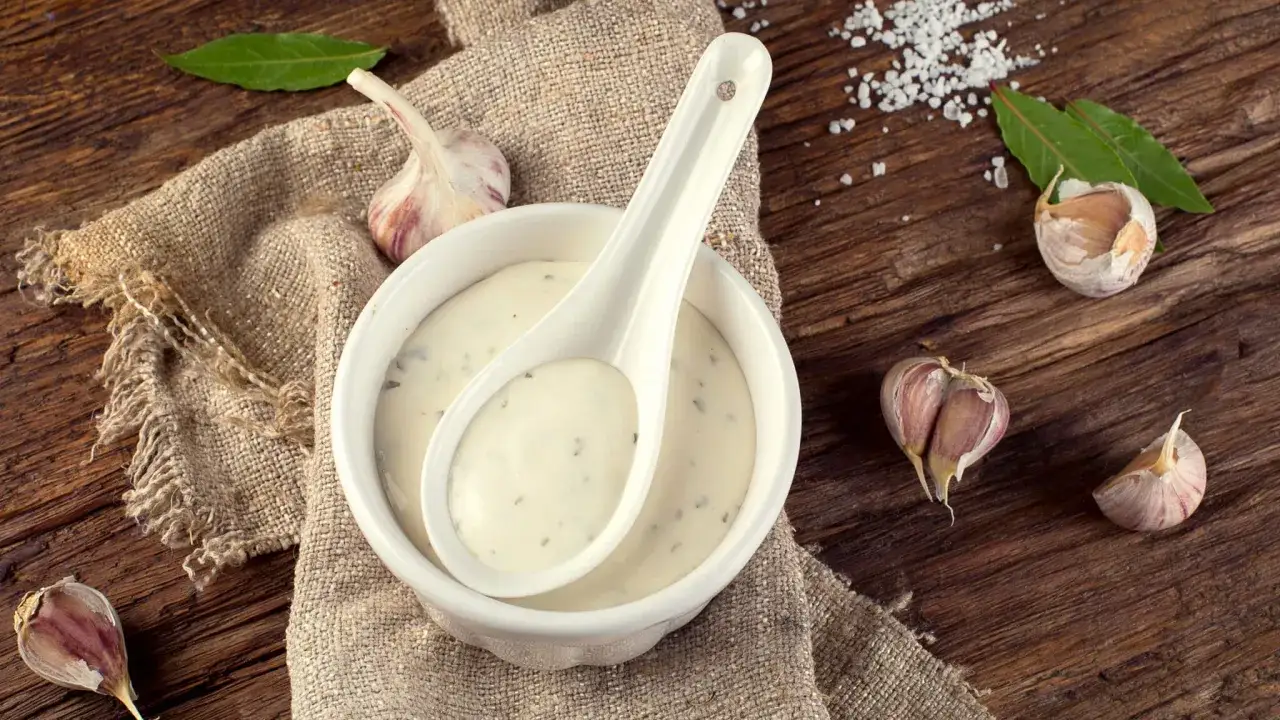 Sauce à l'ail crémeuse dans un bol blanc, avec des gousses d'ail et des feuilles de laurier sur une table en bois rustique.