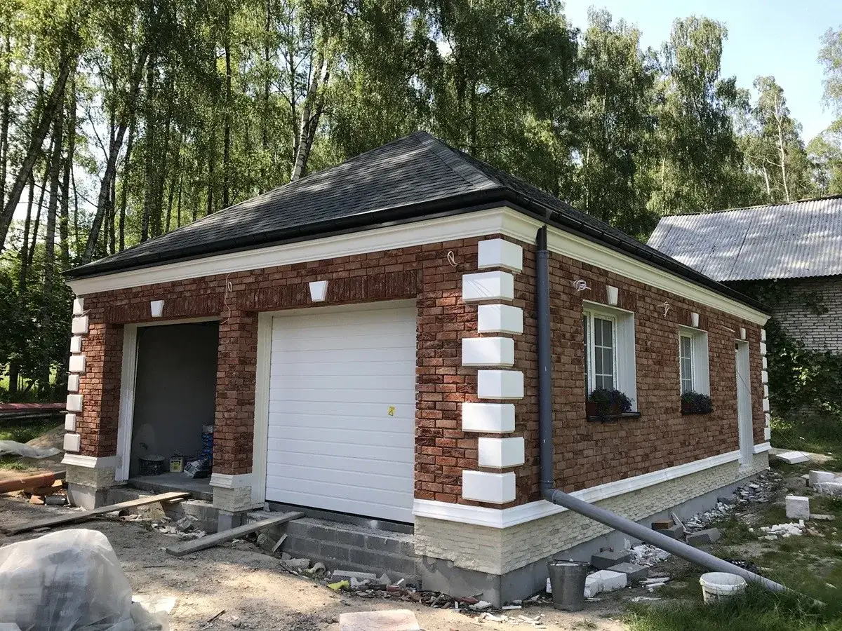 Elegancki garaż z cegły z białym gzymsem nad oknem i drzwiami garażowymi, otoczony zielenią.