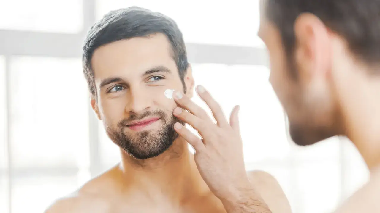 Visage homme 40 ans appliquant de la crème hydratante sur sa joue, reflet dans le miroir.
