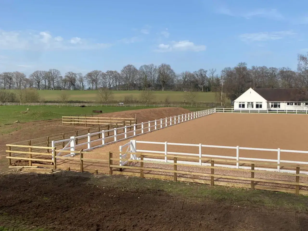 Un grand manège d'équitation avec une piste de sable, entouré de clôtures blanches et en bois. Un bâtiment blanc est visible au fond.