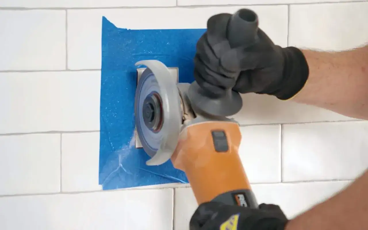 Hands in gloves use an angle grinder to cut a hole in white subway wall tile, demonstrating how to cut wall tile precisely.