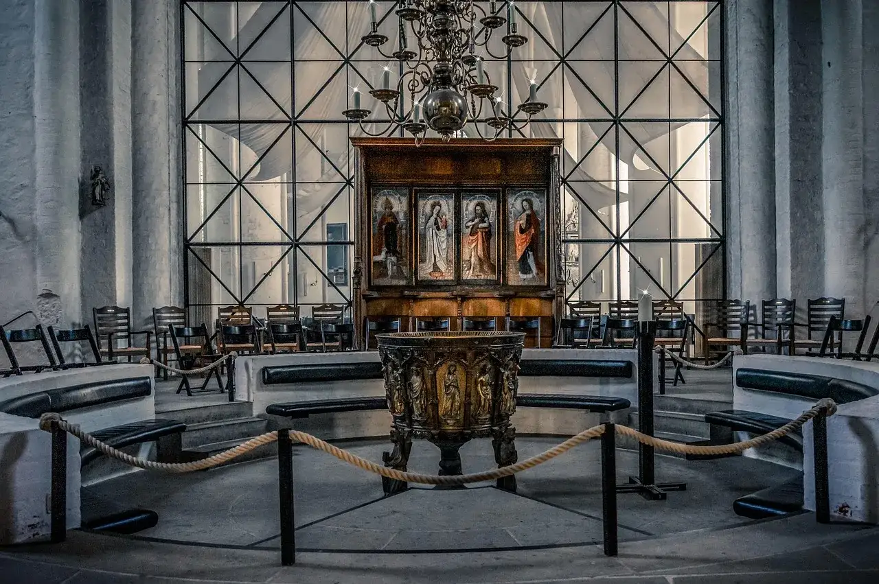 Der katholische Kirche innenraum zeigt einen Taufstein vor einem Altarbild mit Heiligenfiguren. St&uuml;hle und B&auml;nke sind aufgestellt.
