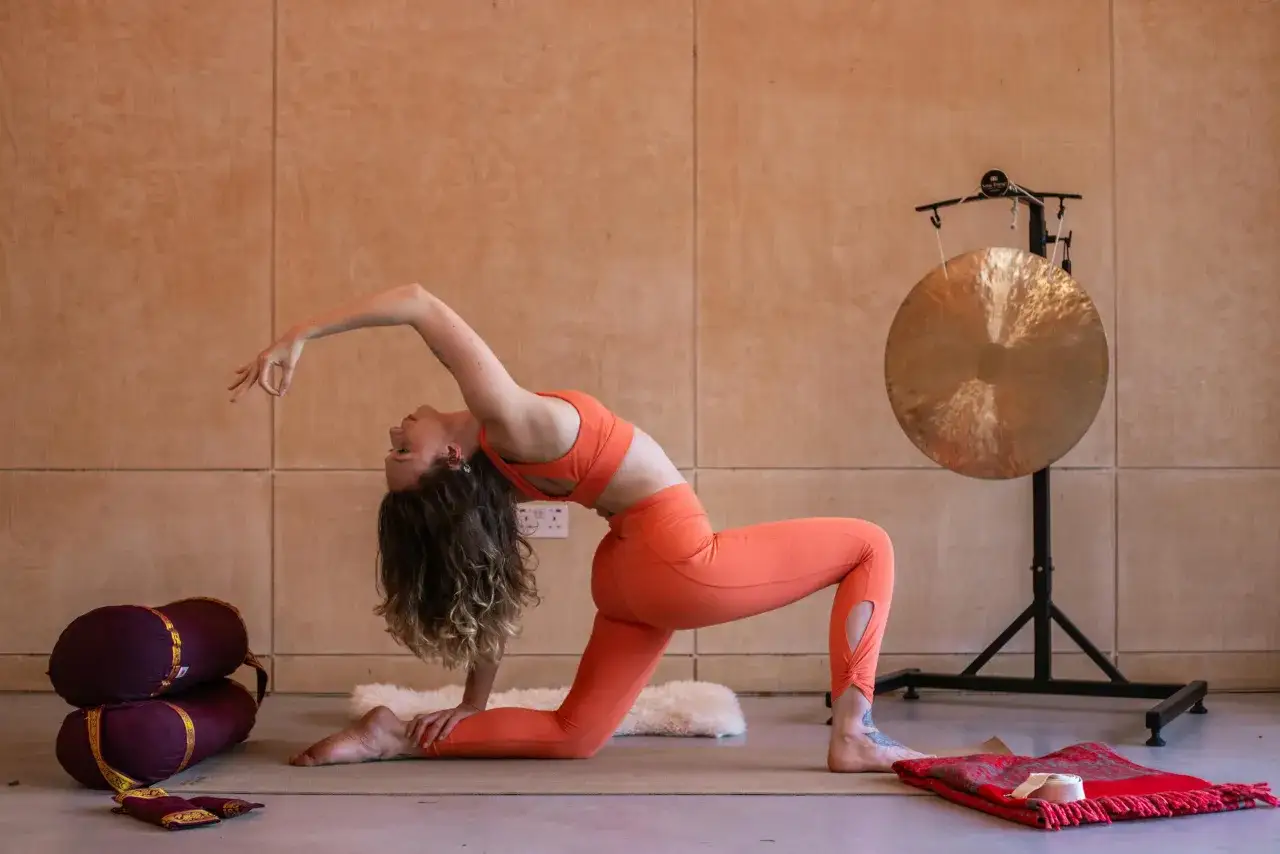 Mujer en postura de yoga Iyengar, con la espalda arqueada y una mano extendida. A su lado, bolsters y un gong.
