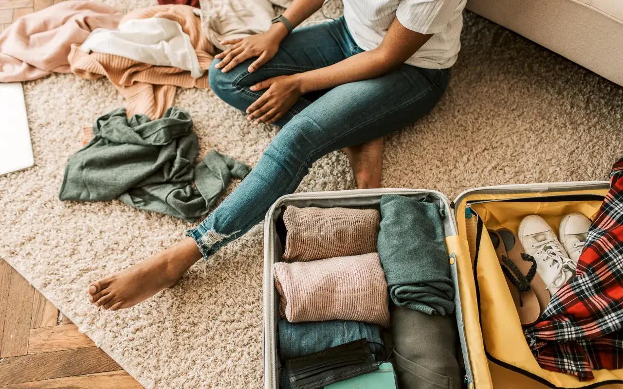 Persona seduta sul tappeto, circondata da vestiti sparsi, con una valigia aperta e quasi piena. L'ansia da viaggio si percepisce nel disordine e nella concentrazione.