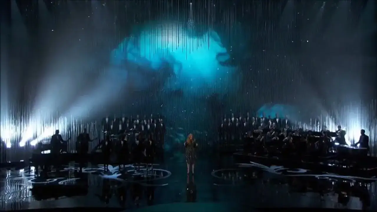 Wokalistka śpiewa Skyfall na tle chóru i orkiestry, w otoczeniu mgły i niebieskich świateł.