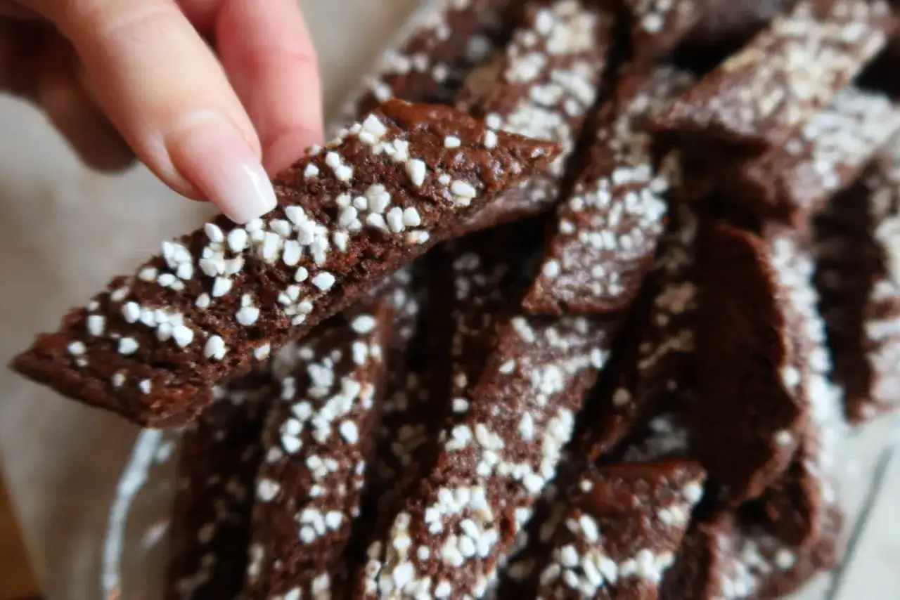 En hand håller en chokladsnitt. Dessa små, mörka kakor är strösslade med vita pärlsocker, perfekta för ett chokladsnittar recept.