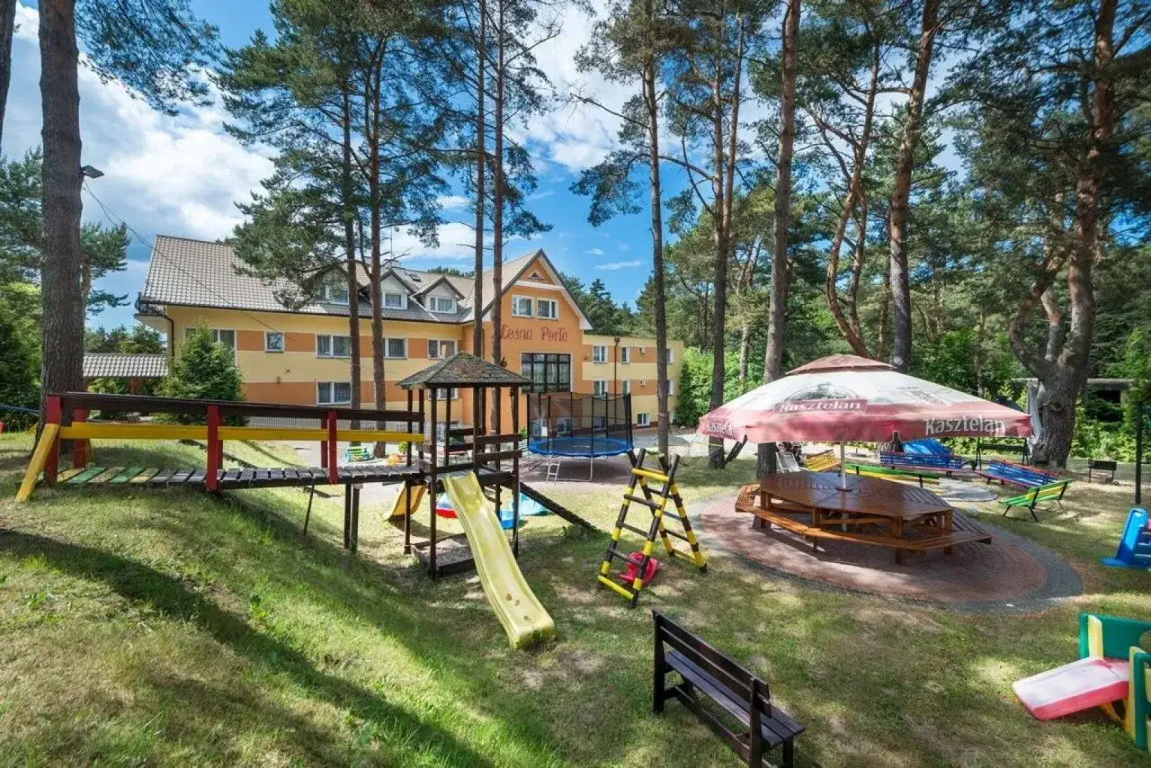 Leśna perła Jastrzębia Góra: plac zabaw z domkiem, zjeżdżalnią i trampoliną, pod parasolem stoły i ławki.