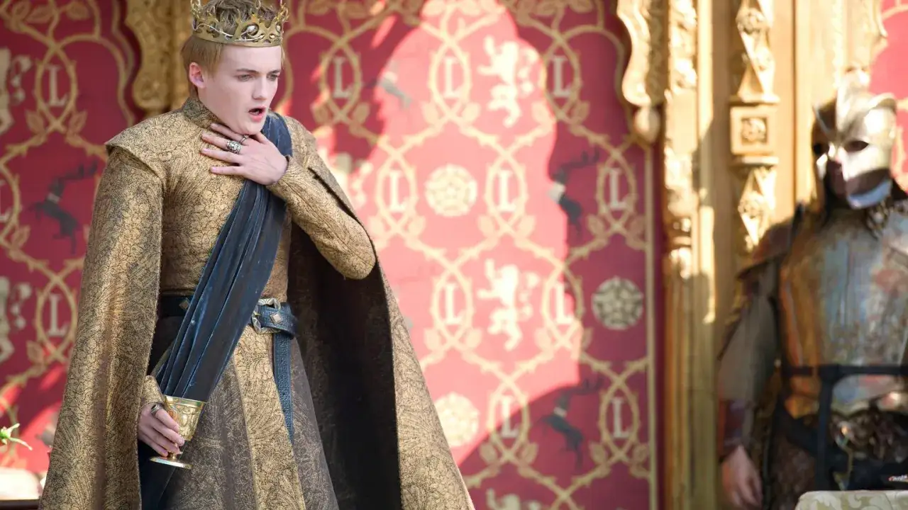Król Joffrey z Gry o Tron, podczas swoich krwawych godów, dławi się winem. Obok stoi rycerz w zbroi.