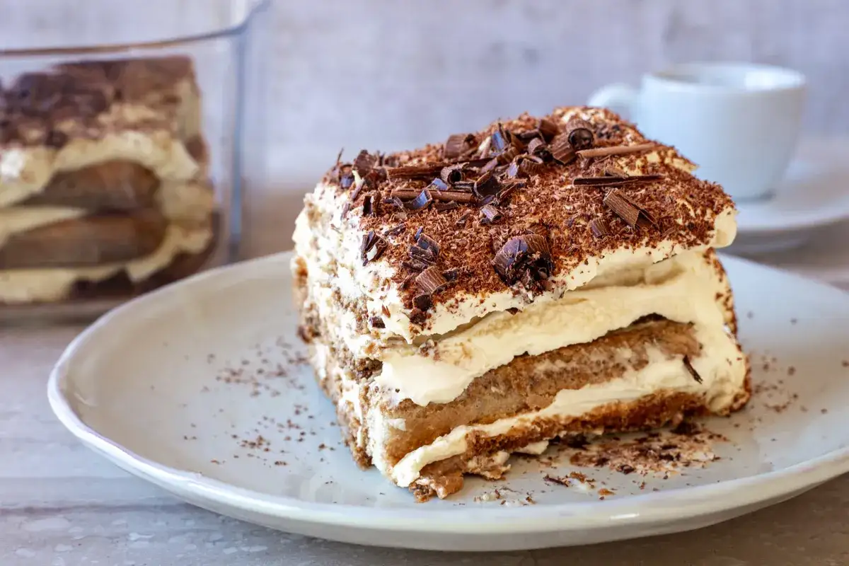Ein Stück Tiramisu, dekorativ mit Schokoladenraspeln bestreut, auf einem Teller. Im Hintergrund ein Glasbehälter mit Tiramisu und eine Kaffeetasse.