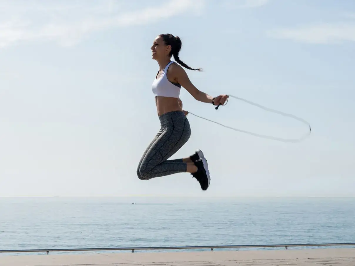 Woman mid-air, enjoying a 10 minute jump rope workout by the ocean. Her braid flies as she leaps.