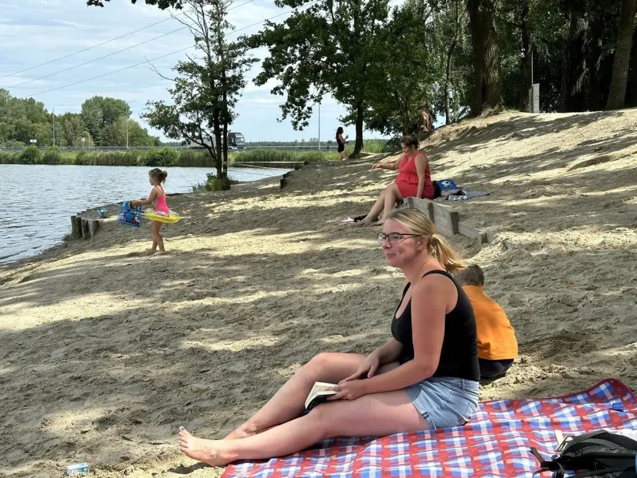 Kobieta czyta książkę na plaży nad jeziorem rybnickim, podczas gdy dziecko bawi się przy brzegu.