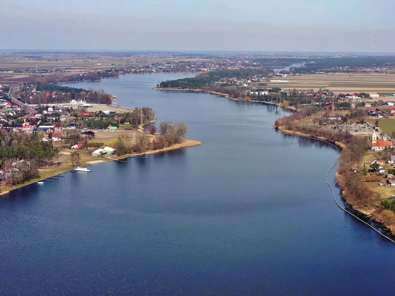 Rozległe jezioro Mikorzyńskie otoczone zabudowaniami i polami. Widoczny kościół i przystań.
