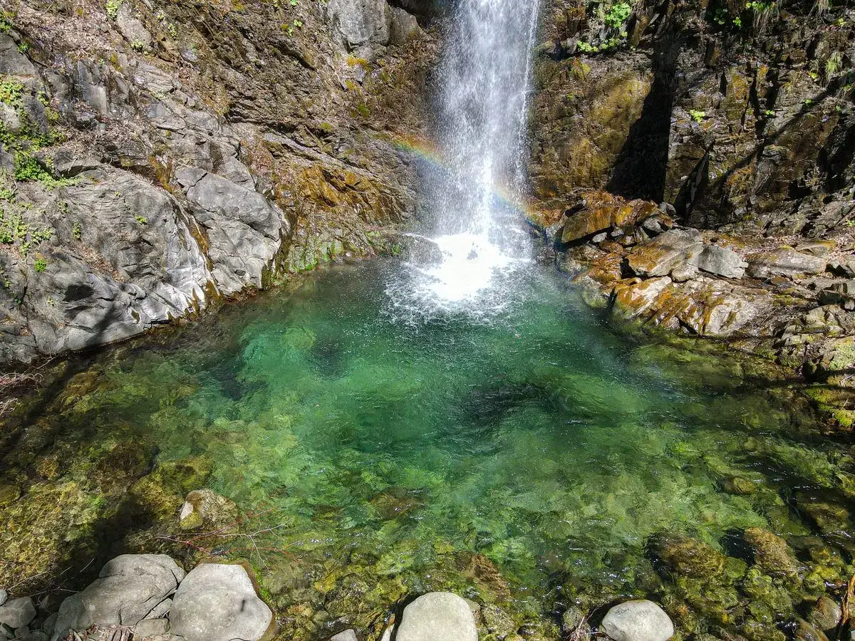 Cascate della Lunigiana: un arcobaleno danza nell'acqua cristallina di una pozza verde smeraldo, incastonata tra rocce scoscese e vegetazione rigogliosa.