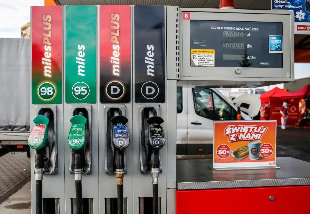 Stacja paliw z dystrybutorami benzyny 98, 95 i **diesel**. Widać pistolety do tankowania, ekran z cenami oraz reklamę hot doga i kawy.
