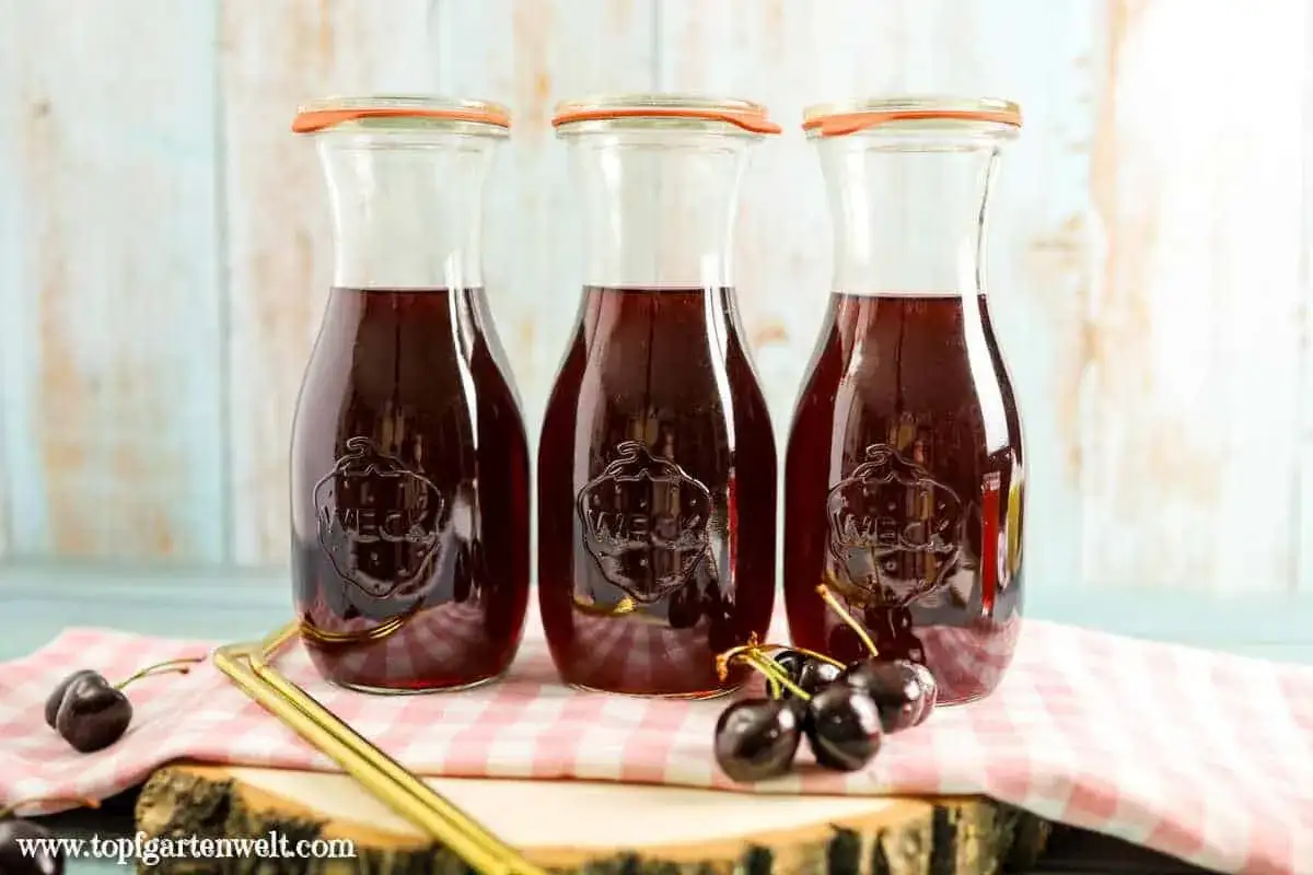 Drei Flaschen selbstgemachter Kirschsaft, bereit zum Genie&szlig;en. Frische Kirschen liegen daneben.
