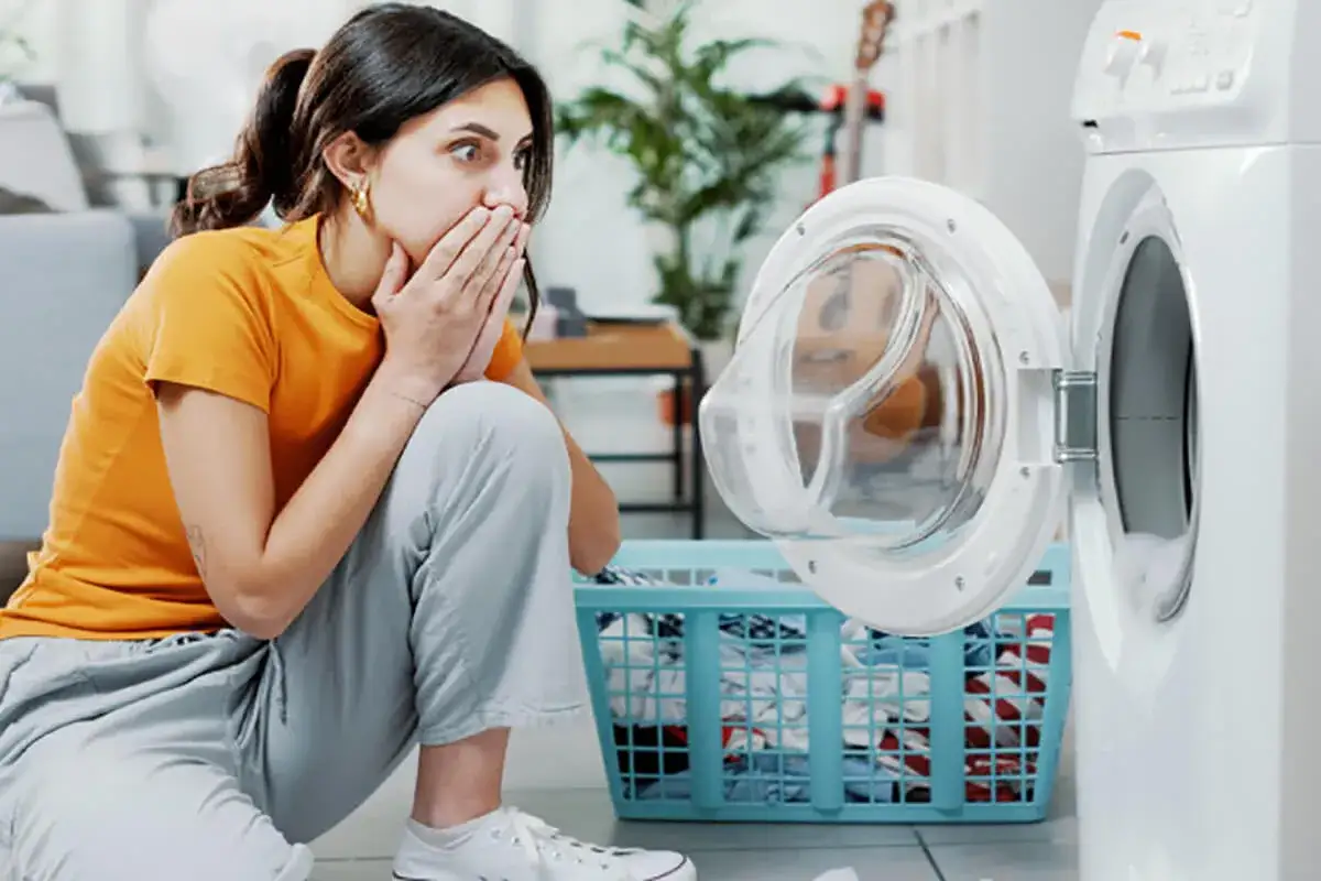 Donna sorpresa con le mani sulla bocca, guarda la lavatrice aperta con un cesto di panni. Forse un ammollo lavatrice inaspettato?