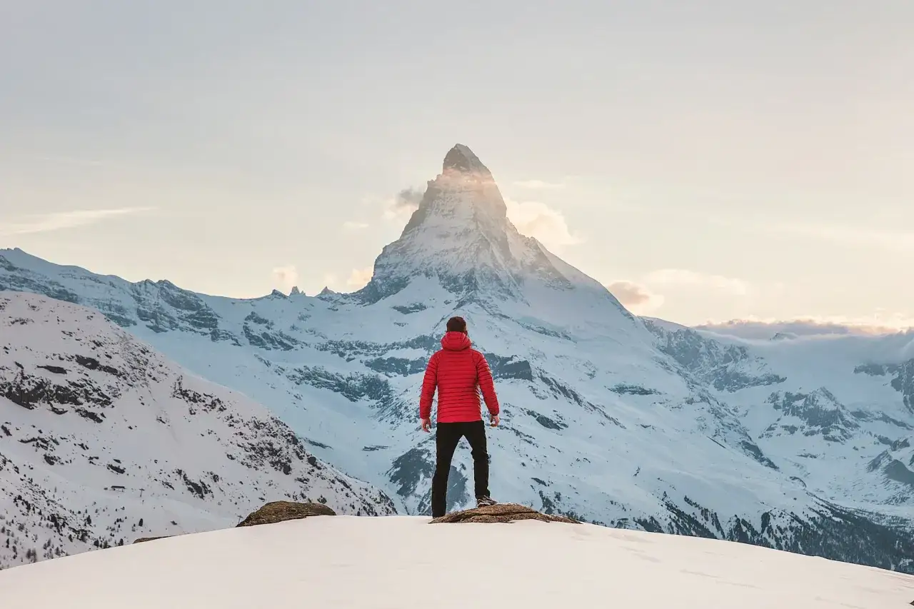 Mann in roter Winterjacke und Hose genießt den Ausblick auf schneebedeckte Berge. Perfekte Kleidung fürs Wandern im Winter.