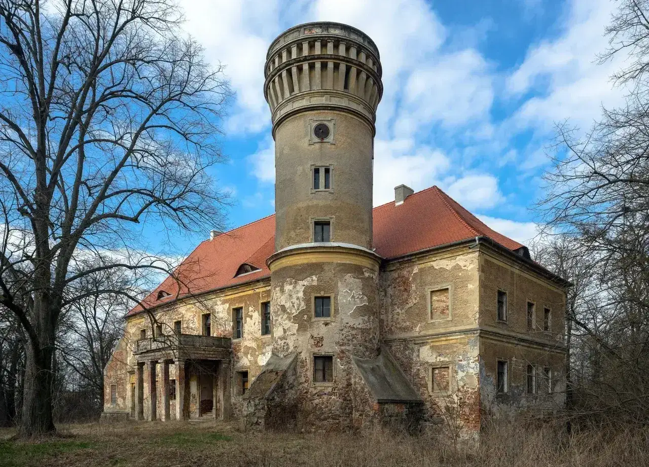 Zniszczony pałac w Osetnie z charakterystyczną wieżą i czerwoną dachówką, otoczony drzewami.