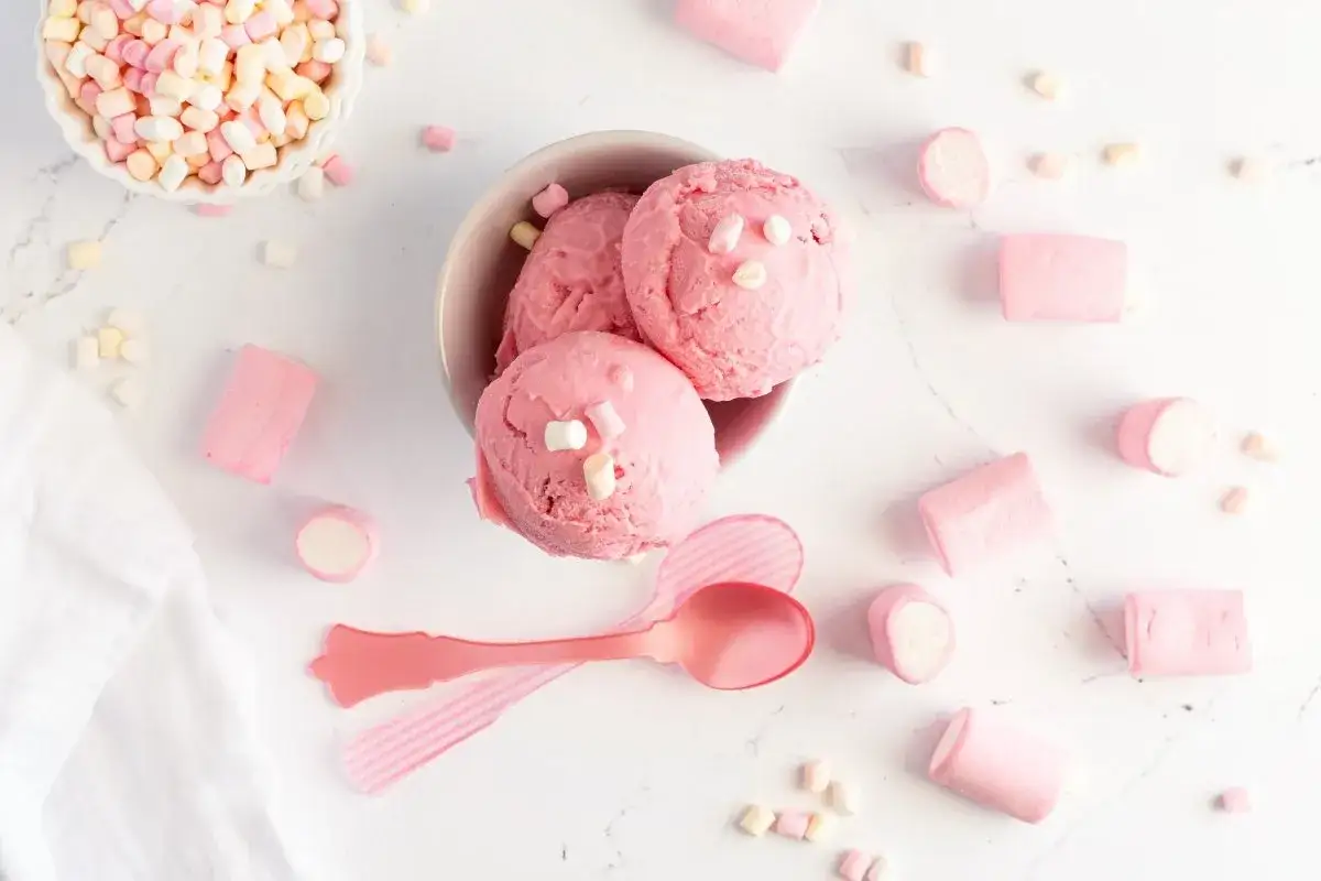 Un delicioso helado de chuche rosa, adornado con malvaviscos, rodeado de más malvaviscos y cucharas rosas.