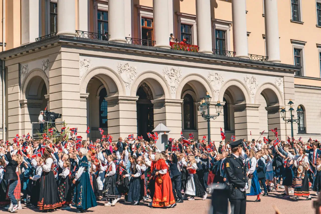 Tłum ludzi, w tym dzieci, świętuje, machając norweskimi flagami. Wiele os&oacute;b nosi tradycyjny str&oacute;j norweski, ozdobiony haftami i kolorowymi wzorami.