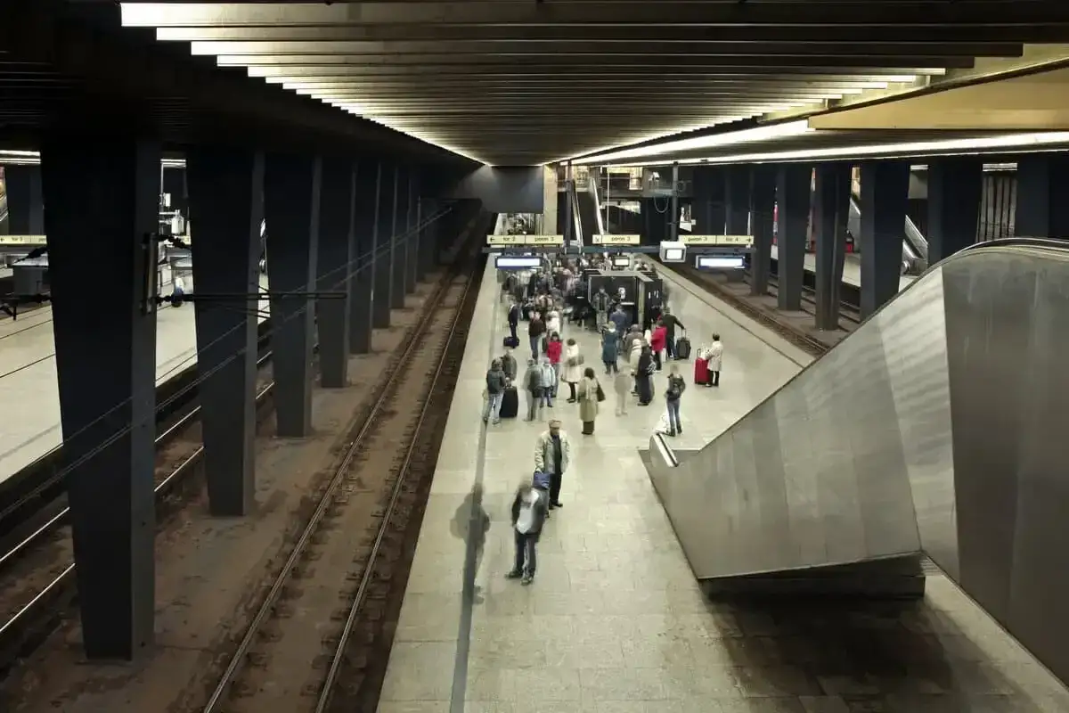 Ludzie na peronie metra, szukający drogi z dworca centralnego do metra. Widać tory, schody ruchome i tablice informacyjne.