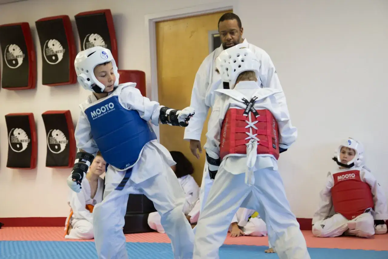 Dzieci w strojach do taekwondo ćwiczą sparing pod okiem trenera. Widoczni są zawodnicy w kaskach i ochraniaczach, gotowi do walki.