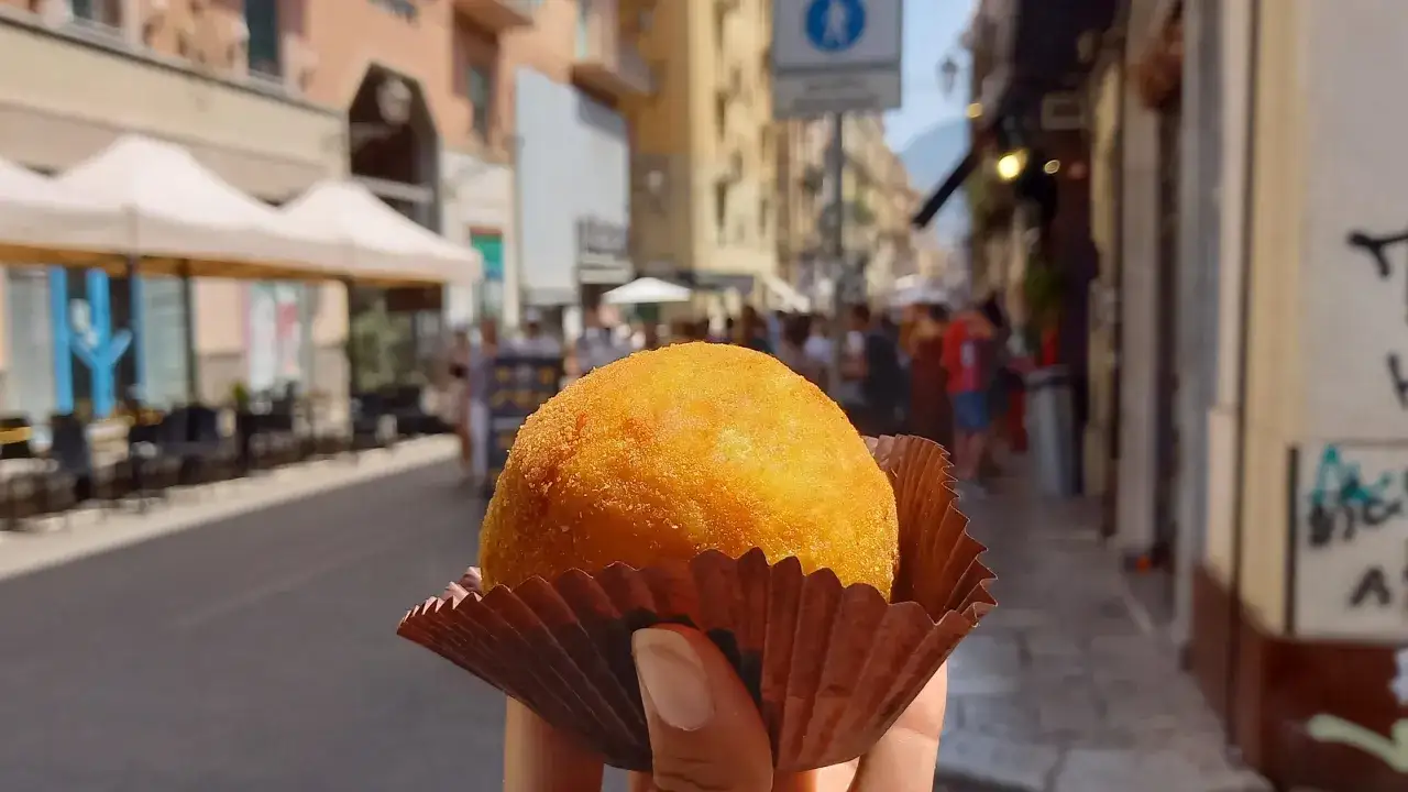 Un arancino dorato, perfetto per un assaggio di **Palermo in un giorno**. La strada animata fa da sfondo a questa delizia siciliana.
