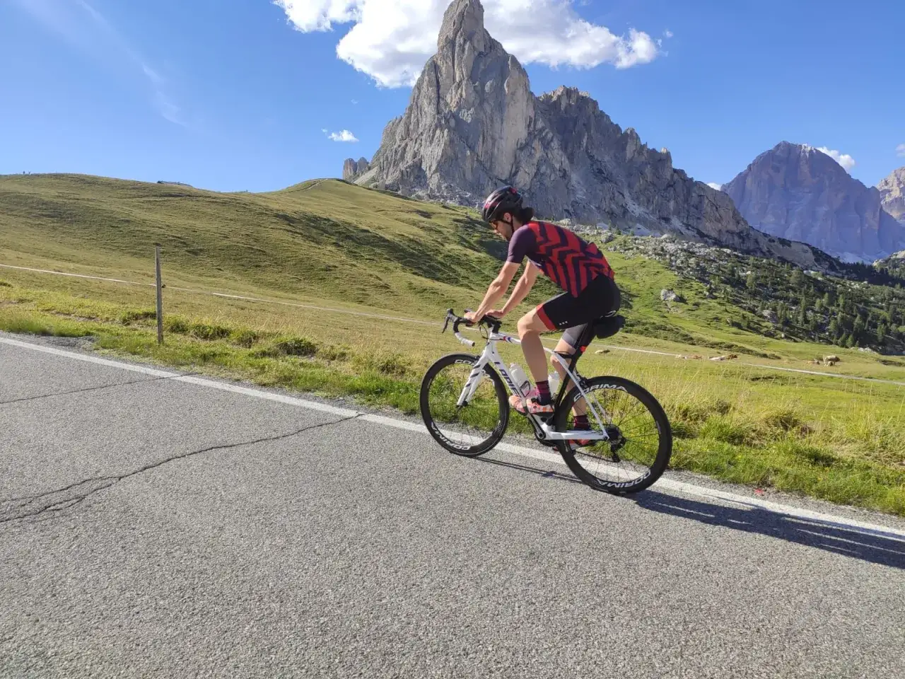 Kolarz na rowerze po włosku pnie się pod górę w Dolomitach. Majestatyczne szczyty i zielone łąki tworzą malowniczy krajobraz.