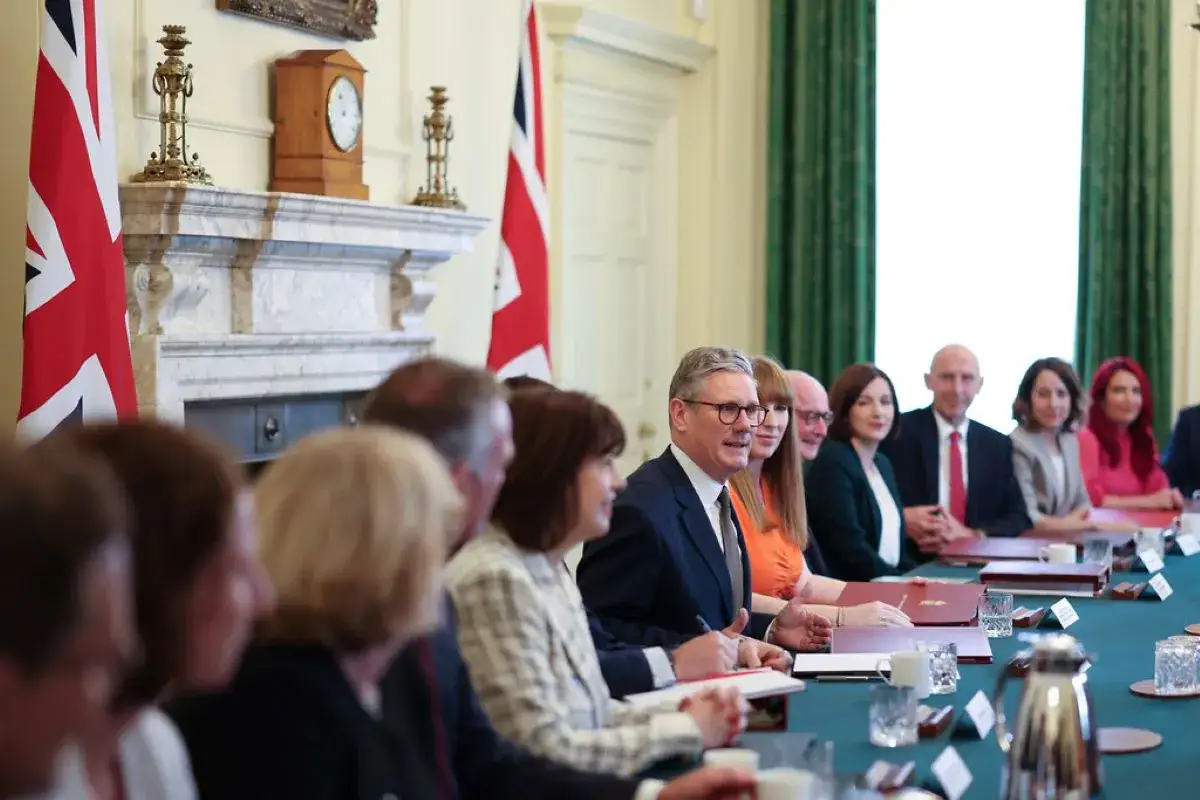 A group of people, including a man who might be a bureaucrat, sit at a long table in a formal room with Union Jack flags.